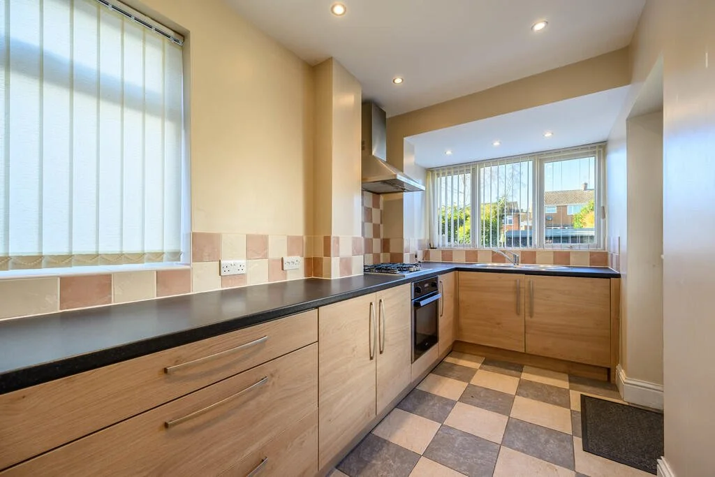 Merritt Estates. Kitchen with wooden cabinets, black countertops, backsplash tiles, and a large window with vertical blinds, showing a suburban neighbourhood outside.