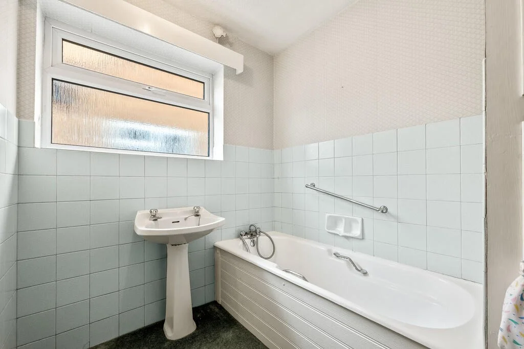 Small bathroom with a tiled wall, a white bathtub with railings, a pedestal sink, and a frosted window.
