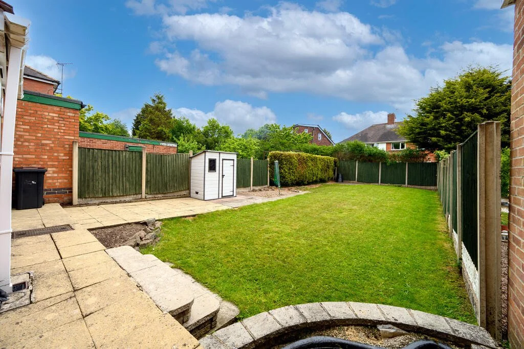 A backyard with a small concrete patio, a gravel path, a green grassy area, a white shed, and a green wooden fence. There are neighboring houses and trees in the background under a partly cloudy sky.