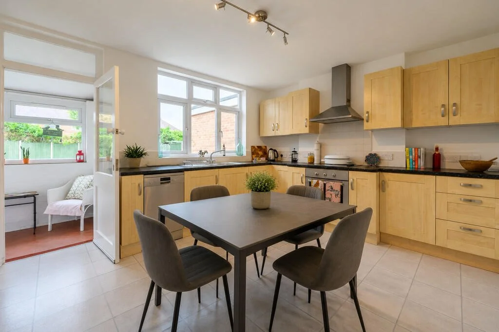 Kitchen with wooden cabinets, black countertop, stainless steel stove and range hood, dining table with four chairs, potted plant on table, window overlooking backyard, small sitting area with chair and side table in sunroom.