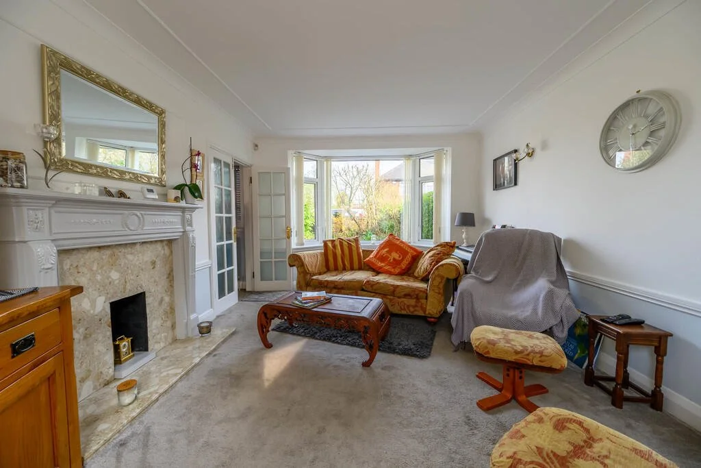 Bright living room with white walls, a fireplace with a gold-framed mirror above it, a large window with bay seating, a floral sofa with throw pillows, an armchair with a grey cover, a wooden coffee table, and various small furniture pieces.