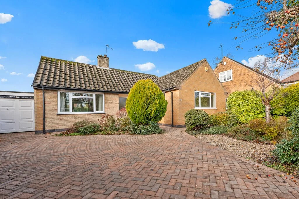 Front view of a detached house with a brick driveway, a garage on the left, and well-maintained shrubs and trees in the front yard.