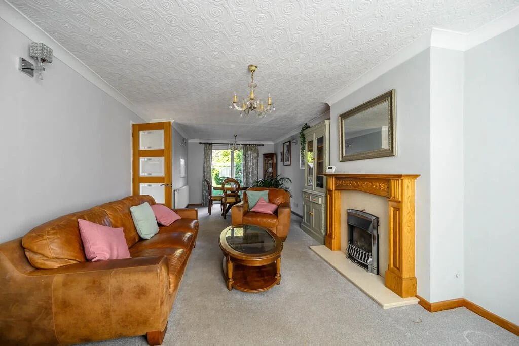 Living room with brown leather sofas, decorative fireplace with wooden mantel, glass-top coffee table, and a dining area in the background with a wooden table and chairs.