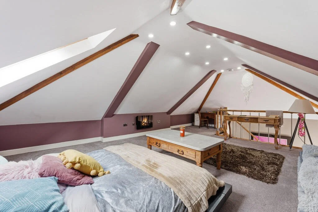 A cozy attic bedroom with sloped ceiling, purple walls, and wooden beams. There is a bed with colorful pillows, a wooden coffee table, and a small fireplace. The space is decorated with a dreamcatcher and a shag rug.