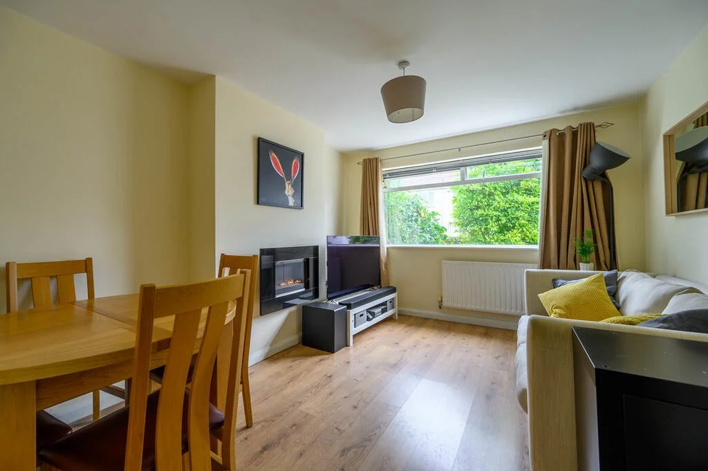 Merritt Estates. Living room with a beige sofa with yellow and grey pillows, a window with beige curtains, a wooden dining table with chairs, a television, a fireplace, and an art piece of a rabbit on the wall.