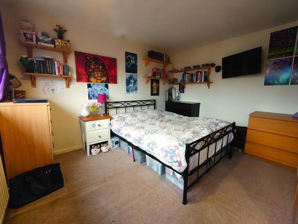 A bedroom with a bed, black metal frame, and a quilt. There are wooden shelves with books, artwork, and decorative items on beige walls. Small furniture pieces include a white nightstand with a pink lamp, a black dresser, and a wooden dresser. A TV is mounted on the wall, and a flat-screen monitor is on the dresser. There is a black bag on the beige carpeted floor.
