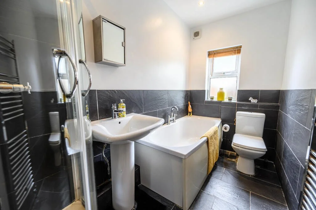 Merritt Estates. A small bathroom with a bathtub, sink, toilet, and window with blinds. The walls are partially tiled in dark grey, and the floor is tiled in matching dark grey. The bathroom has a minimalist design with a few decorations.