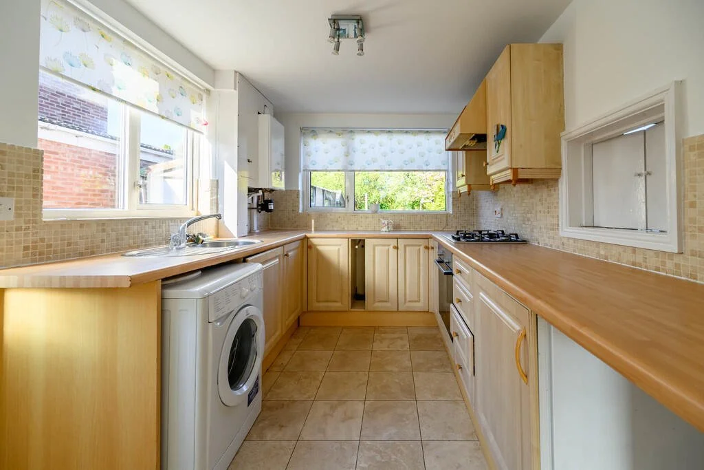 A bright kitchen with wooden cabinetry, beige tile flooring, and a beige tiled backsplash. There are three windows with floral curtains letting in natural light. Appliances include a washing machine under the counter, a gas stove, and an oven. The countertops are wooden.