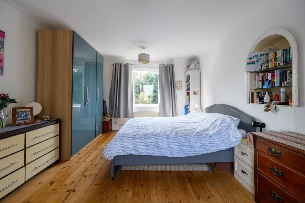 A bedroom with a bed, a window with curtains, a white bookshelf, a large wardrobe, and a wooden dresser.