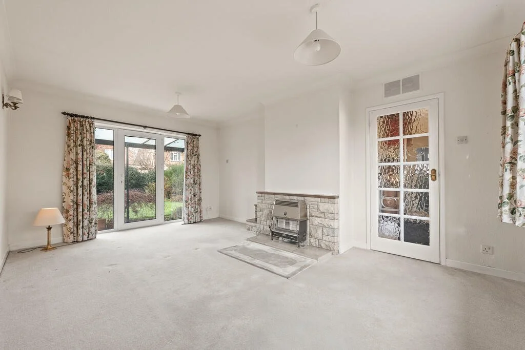 Living room with large glass doors opening to garden, floral curtains, a small lamp on the carpet, a fireplace with stone surround, and a door with textured glass panels.