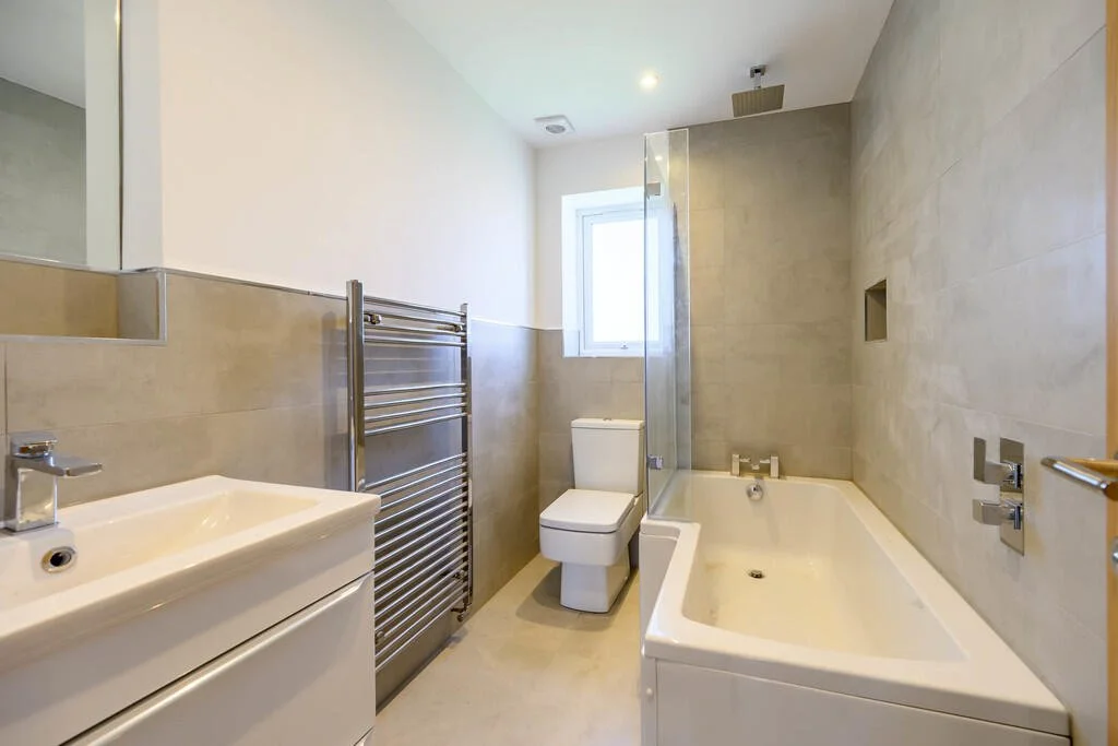 Modern bathroom with beige tiled walls, white bathtub, toilet, small sink, and towel heater.