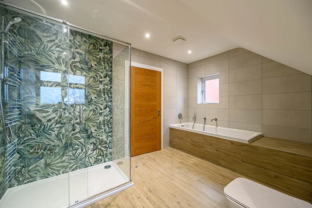 Merritt Estates. Modern bathroom with walk-in shower featuring tropical leaf-patterned glass, a wooden door, built-in bathtub, small window, and wooden flooring.