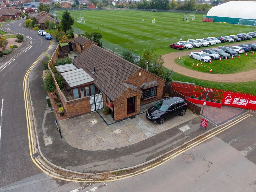 Merritt Estates. An aerial view of a brick house with a small driveway and a parked black car, adjacent to a sports field with goal posts and surrounded by a parking lot and a sign for The Nigel Doughty Academy.