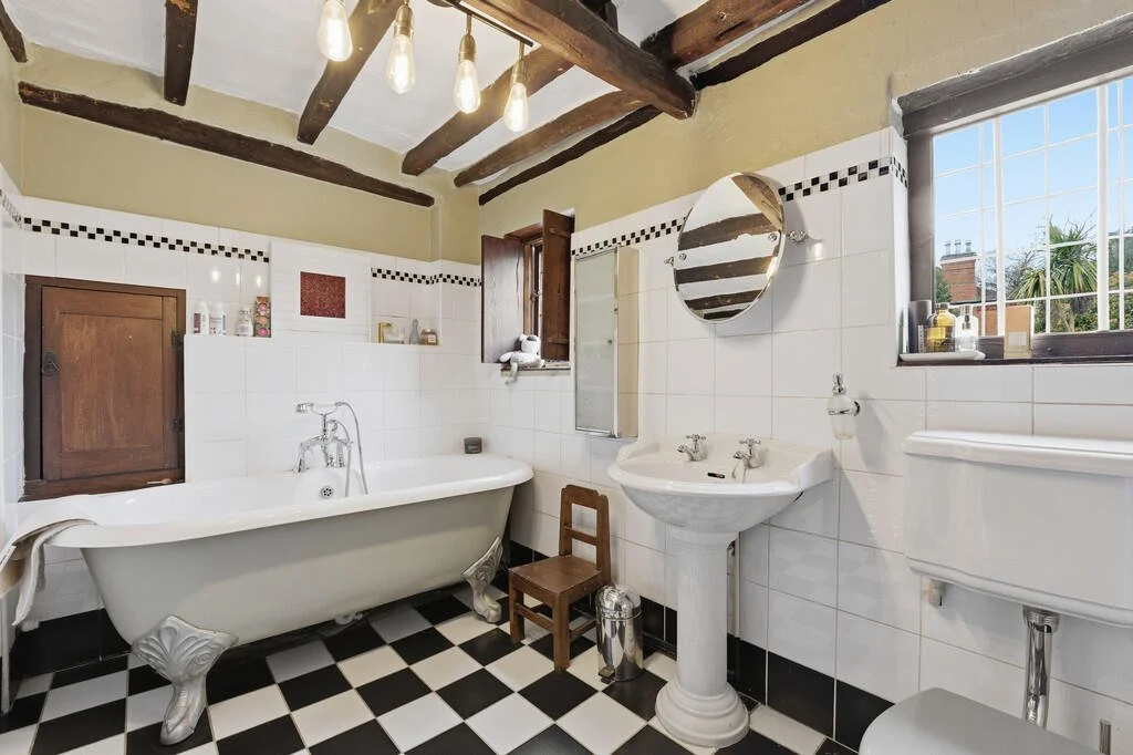 Bathroom with vintage clawfoot bathtub, pedestal sink, and toilet, featuring black and white checkered floor tiles, wooden ceiling beams, and a window with a view of trees outside.