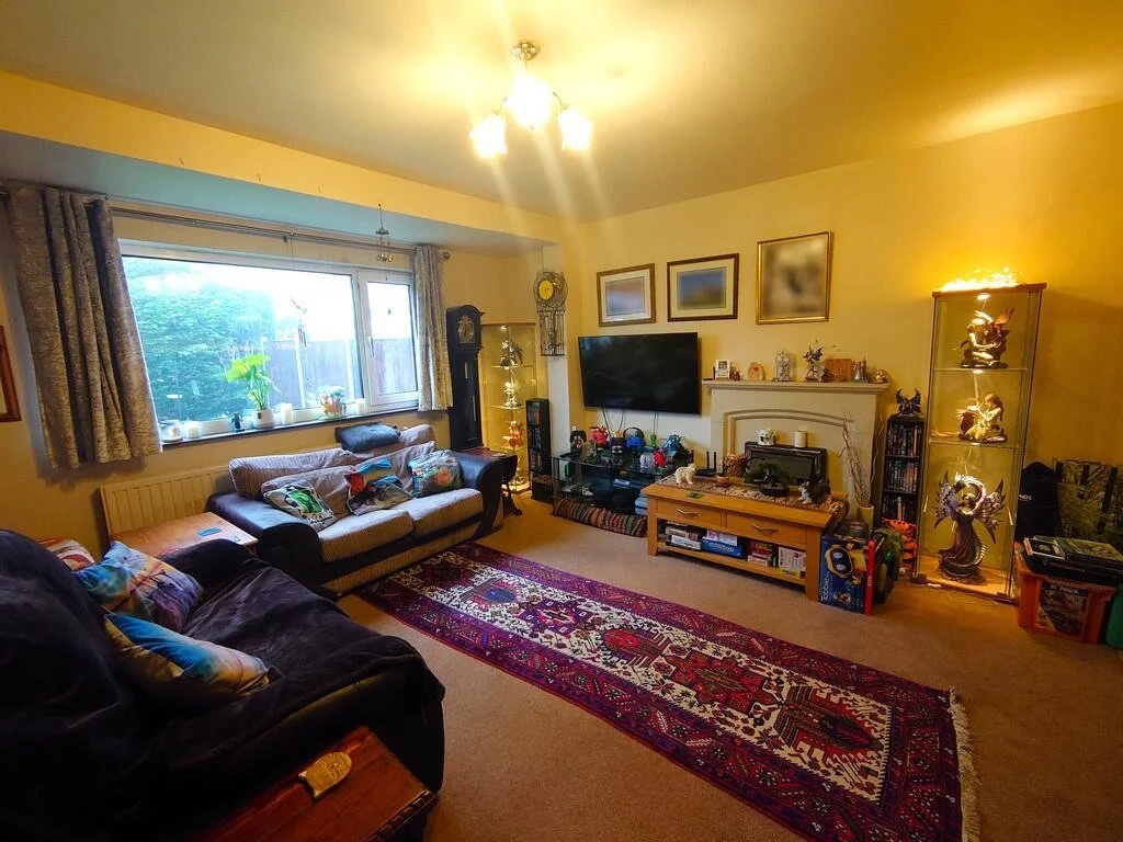 A cozy living room with a large window, beige walls, and a colorful patterned rug. There are two sofas with decorative pillows, a television on a stand, a tall corner display case with collectibles, and wall art. The room is illuminated by ceiling lights.