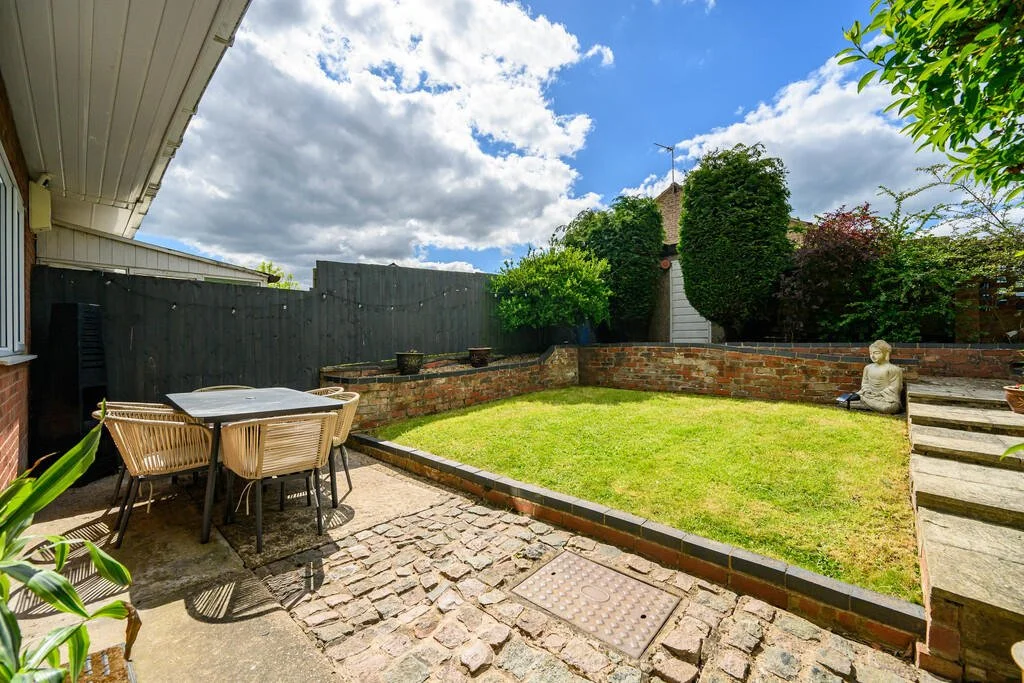 A backyard patio with a table and chairs on a cobblestone surface, a small grass patch surrounded by brick borders, a statue of Buddha, lush greenery, and a cloudy blue sky.