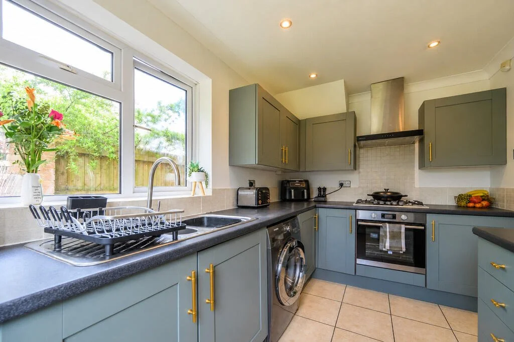 Kitchen with grey cabinets, a black countertop, a washing machine, a stove, and a window with a view of green trees and a wooden fence.