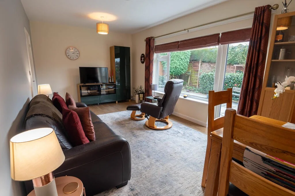 Merritt Estates. Living room with a black leather sofa, a floor lamp, a TV on a stand, a recliner chair facing the window, and a large window with brown curtains overlooking a garden.