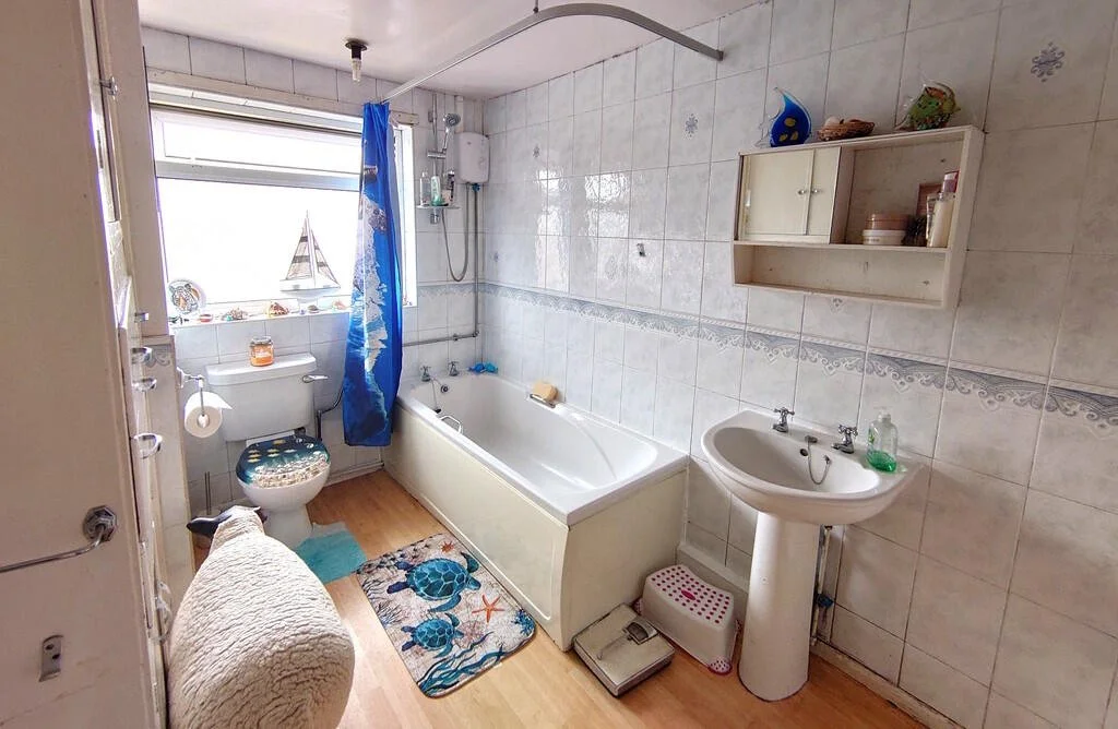 A bathroom with a bathtub, a toilet with a turtle-patterned toilet seat cover, a small window with a sailboat decoration, and a white sink. There is a blue shower curtain, a wooden floor, and a white tile wall with decorative border. Items on shelves include a blue ball, bowls, and toiletries.