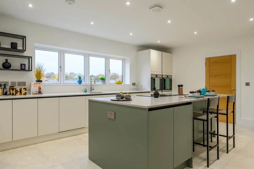 Merritt Estates. Modern kitchen with white cabinetry, an island with a green base, a window with a view outside, and black chairs at the island with teal tableware.