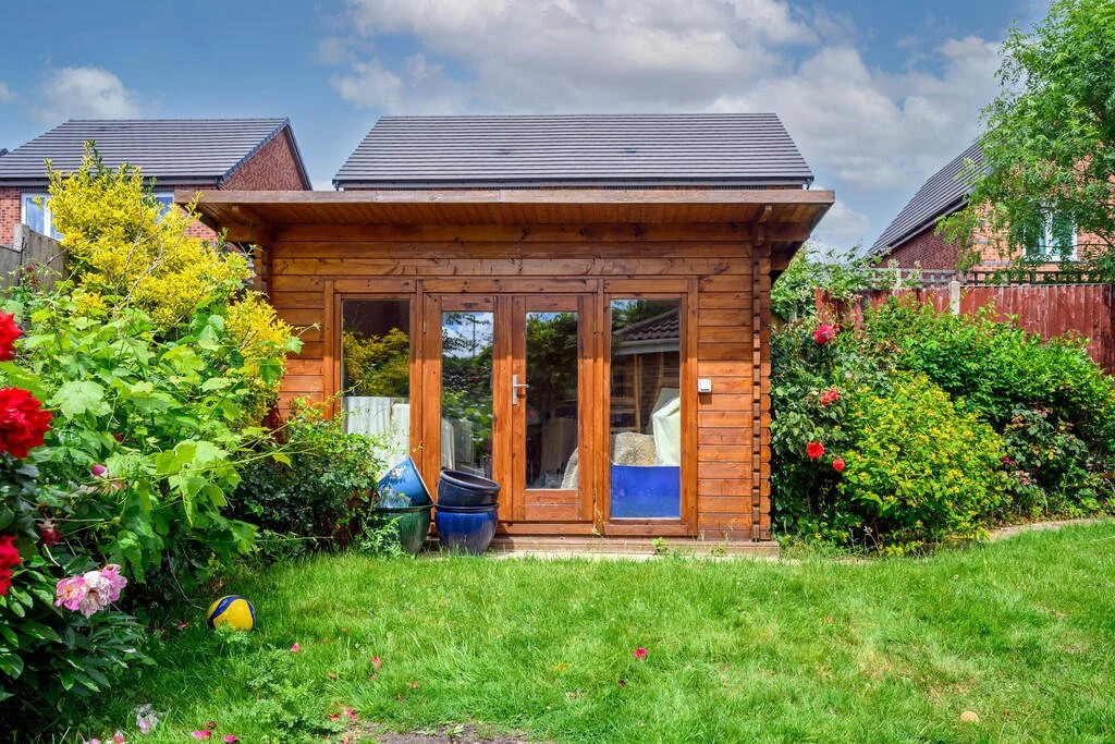 A wooden garden shed with glass doors, surrounded by lush green bushes and flowering plants, in a suburban backyard with neighboring houses visible in the background.