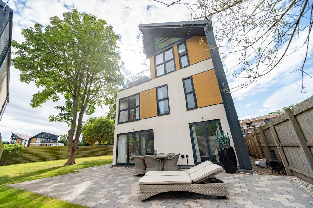 Merritt Estates. Modern multi-story house with a backyard patio, outdoor furniture, and a large tree. The house has a mix of white and wooden panel exterior, with large windows and sliding glass doors.