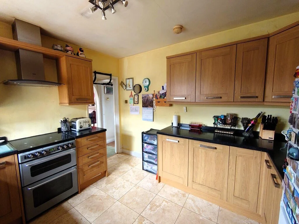 Kitchen with wooden cabinets, black countertops, stainless steel oven, toaster, and various small kitchen items, beige tiled floor, yellow walls, and a door leading to another room.