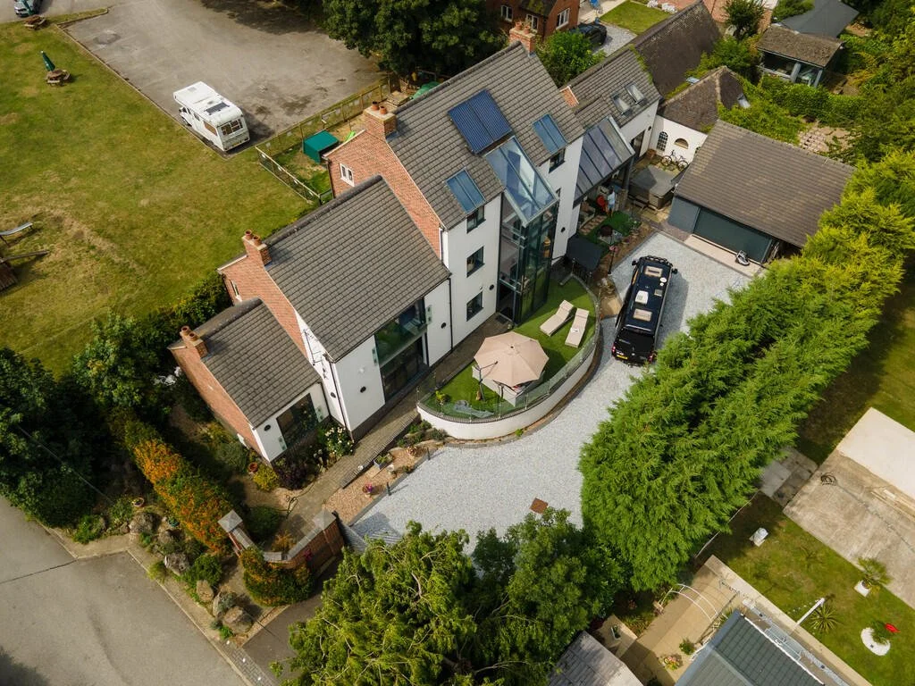 Merritt Estate. Aerial view of a modern multi-story house with a glass-enclosed staircase, a gravel driveway with a black vehicle, a small backyard with an umbrella, lounge chairs, and garden furniture, surrounded by green trees.