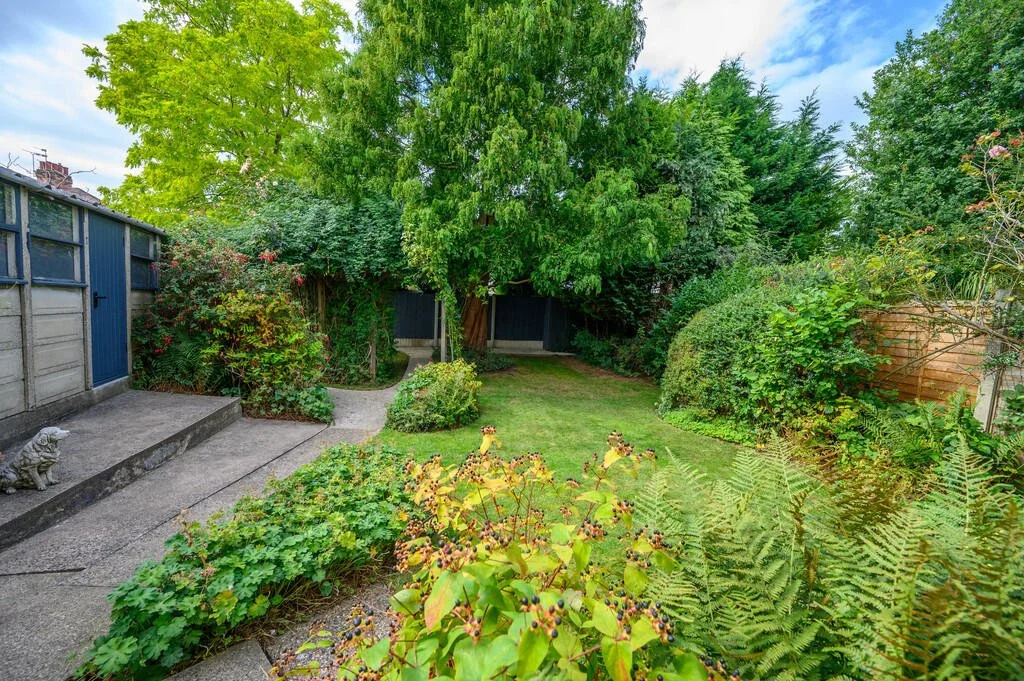 Merritt Estates. A lush green backyard with a variety of trees, bushes, and ferns, featuring a grassy lawn area, a concrete patio with a small statue of a dog, and a wooden fence in the background.
