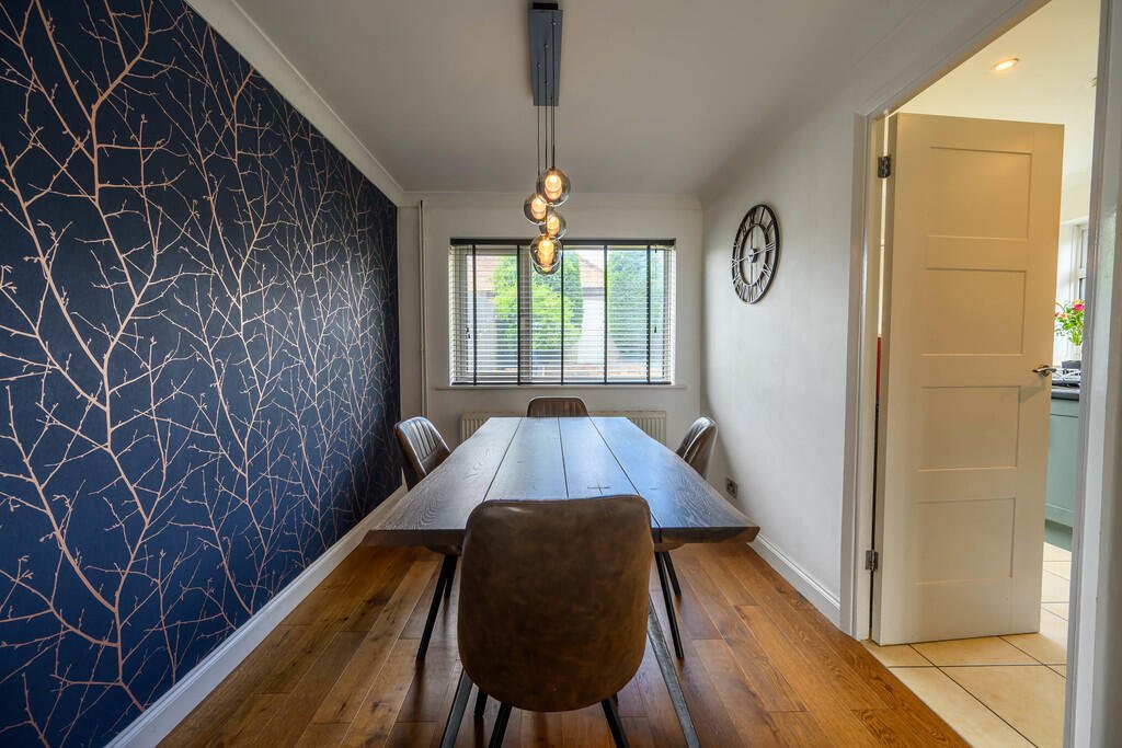 Dining room with a wooden table, four chairs, a wall clock, and a window with blinds, with a navy accent wall featuring a gold leaf pattern.