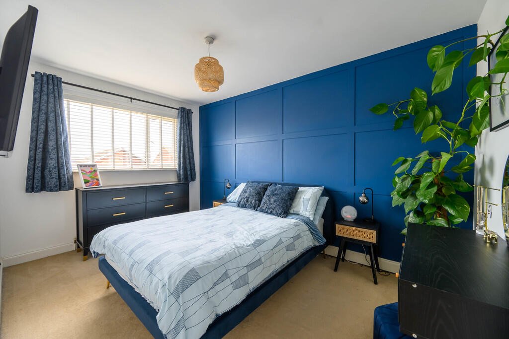 A bedroom with a blue accent wall, a bed with white and blue bedding, black side tables, a window with blue curtains, a black dresser, a TV, and a large green plant.