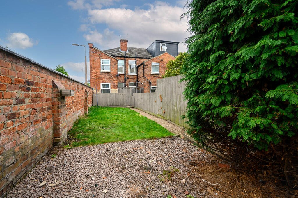 Merritt Estates. Small backyard with a gravel path, a strip of grass, a brick wall on the left, a wooden fence at the back, and a large green bush on the right, with multi-story brick houses in the background under a partly cloudy sky.