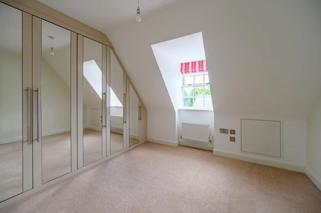 Empty attic bedroom with beige carpet, built-in mirrored wardrobe, and window with pink-striped Roman shade.