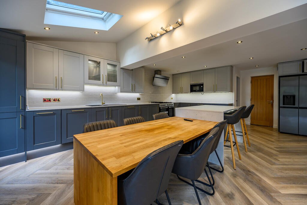 Merritt Estates. Modern kitchen with grey and blue cabinets, a large wooden dining table with black and grey chairs, a skylight, and hardwood flooring.
