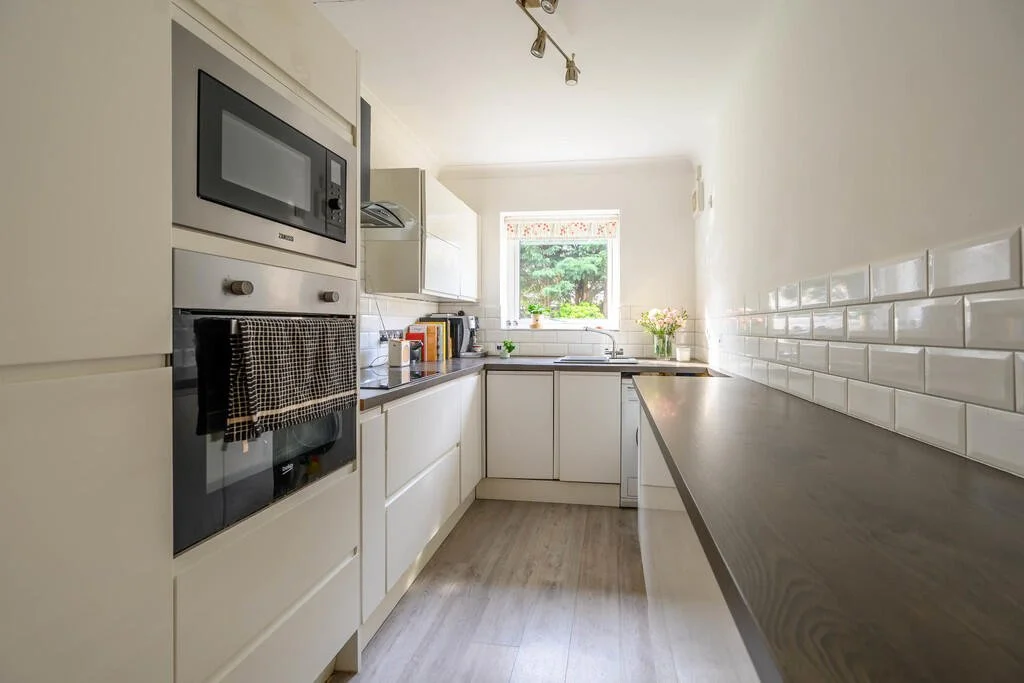 Small, modern kitchen with white cabinets, a built-in microwave, an oven with a towel, a window with a view of greenery, flowers on the counter, and white subway tile backsplash.