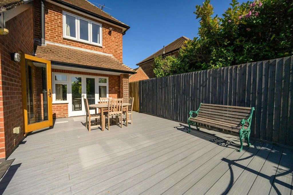 A spacious backyard deck with outdoor furniture including a wooden table and chairs, a green metal swing bench, a wooden privacy fence, and the back of a brick house with a sliding glass door and windows.