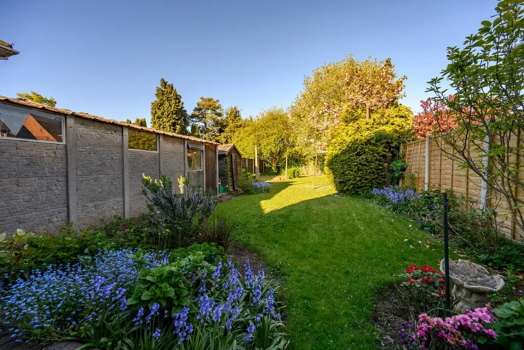 A backyard garden with a lawn, colorful flowers, trees, and a wooden fence on a sunny day.