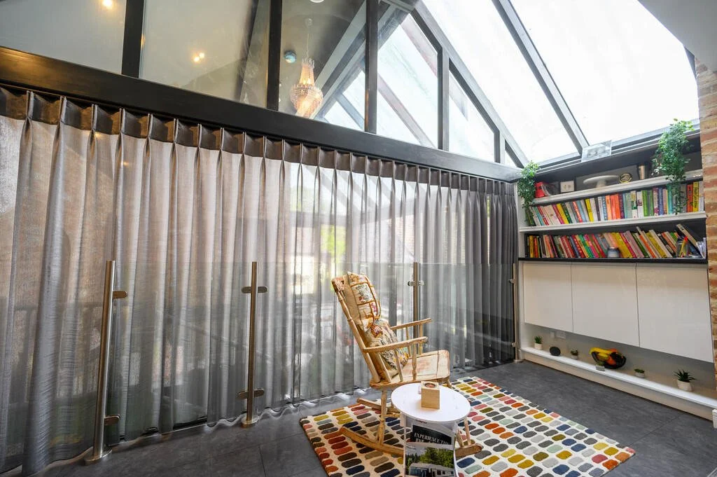Merritt Estates. A cosy living space with a rocking chair, small round white table, colourful patterned rug, tall curtains, bookshelf filled with colourful books, and large windows with triangular glass panes letting in natural light.