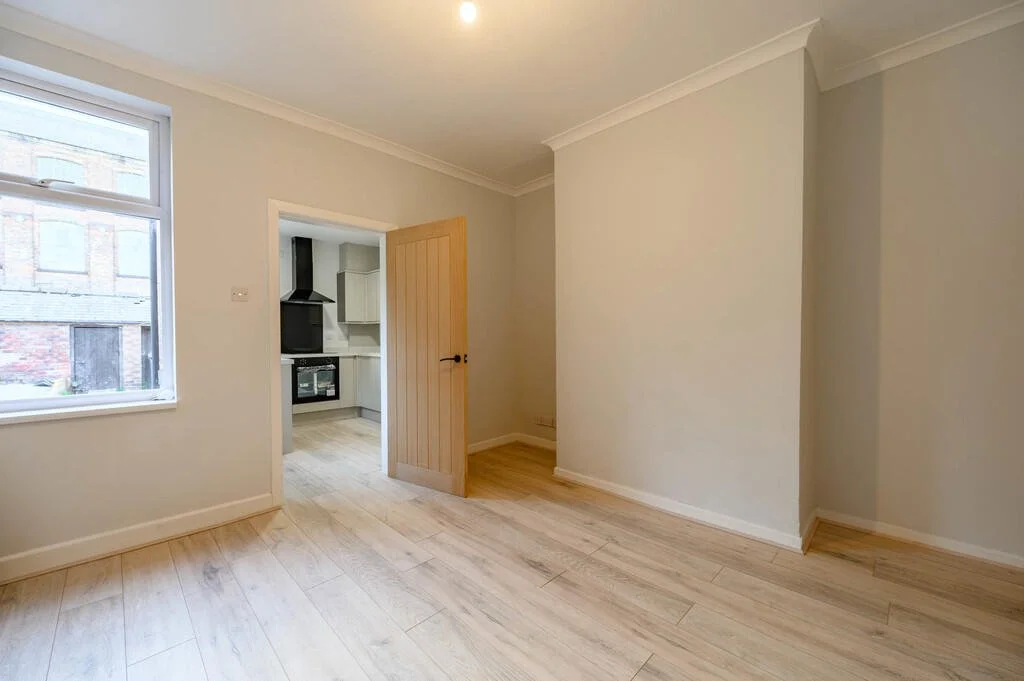 Merritt Estates. Empty room with cream-colored walls, light wood flooring, and a window revealing an outside brick building. An open wooden door leads to a kitchen with white cabinets and black oven range.