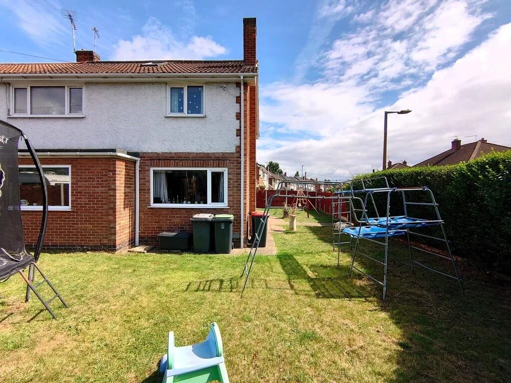 A backyard with a two-story house, green grass, a trampoline on the left, a small slide in front, and climbing frames on the right side. The house has a brick lower floor and a white upper floor, with sliding glass doors and windows. There are trash bins outside, and the sky is blue with some clouds.
