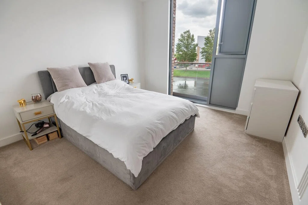 A minimalist bedroom with a bed, two bedside tables, and a large window with a balcony door, overlooking green trees and a cloudy sky.