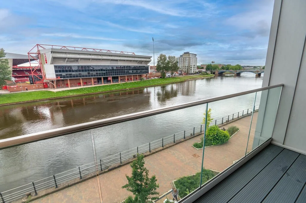 View from a balcony overlooking a river with a modern stadium on the opposite bank, trees, a walkway, and a bridge in the background under a cloudy sky.