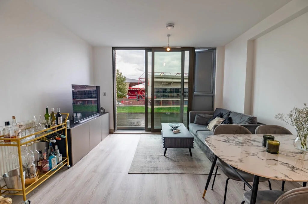 Modern living room with a gray couch, coffee table, flat-screen TV, and sliding glass doors leading to a balcony with a view of a sports stadium.
