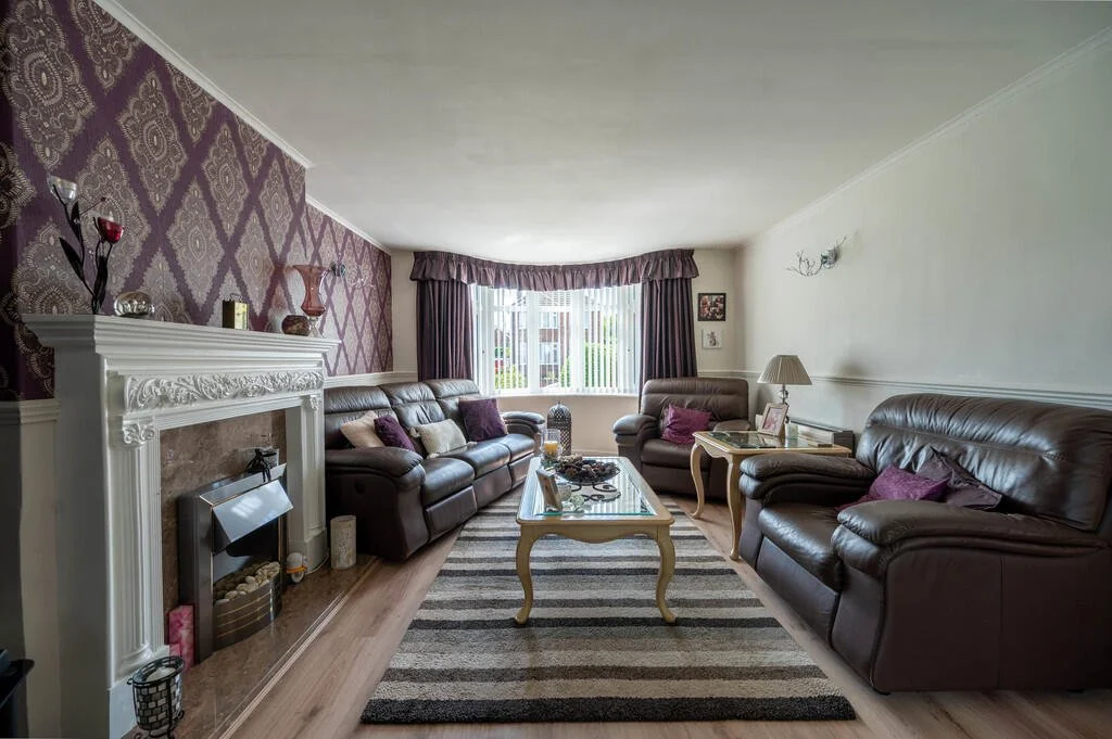 Living room with purple and white patterned accent wall, leather sofas, a striped rug, a fireplace with decorative items, a center coffee table, side tables with lamps, window with curtains, and a few wall decorations.