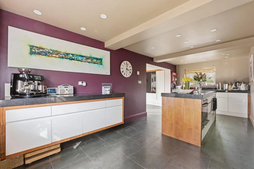 Modern kitchen with white cabinets, black countertops, and a purple accent wall featuring a colorful abstract painting and a large clock, with additional countertops and appliances in the background.