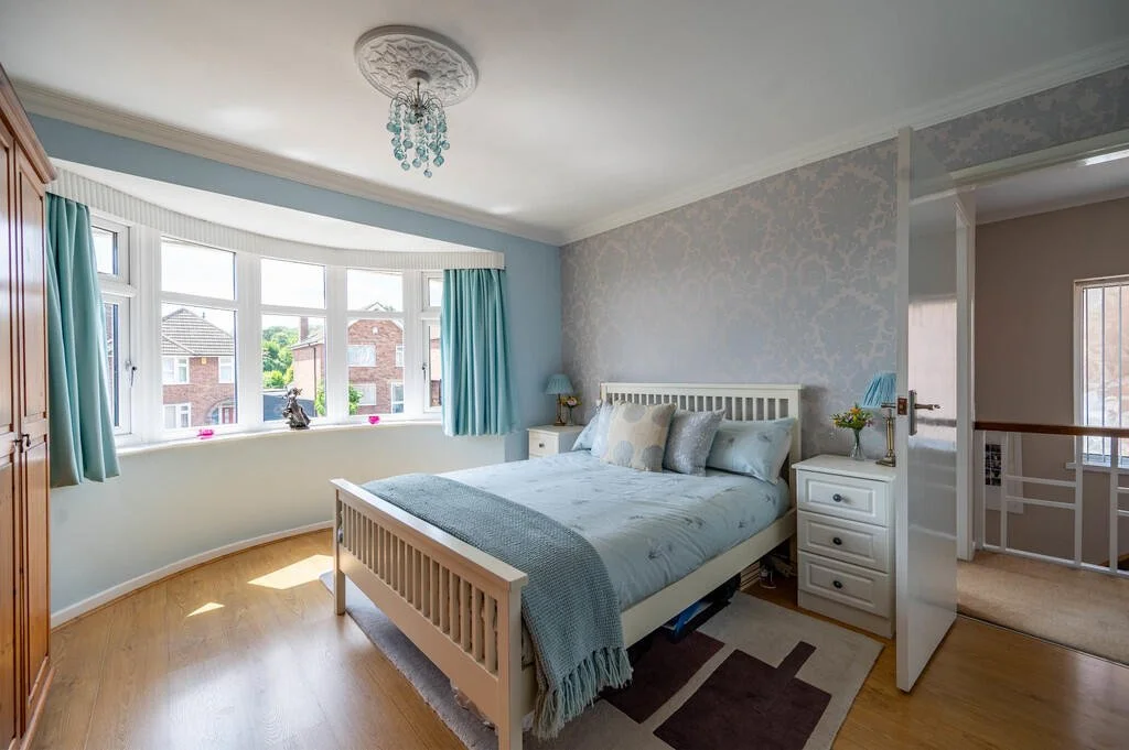 A cozy bedroom with light blue walls, a white bed with multiple pillows, a small white nightstand with a lamp and flowers, a large bay window with curtains, a wooden wardrobe, and hardwood floors.