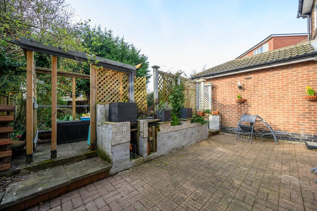 A small backyard patio with brick paving, a couple of garden chairs, potted plants on a brick wall, and a raised garden bed with greenery. There is a wooden fence and latticework at the back, and part of a house with a brick exterior.