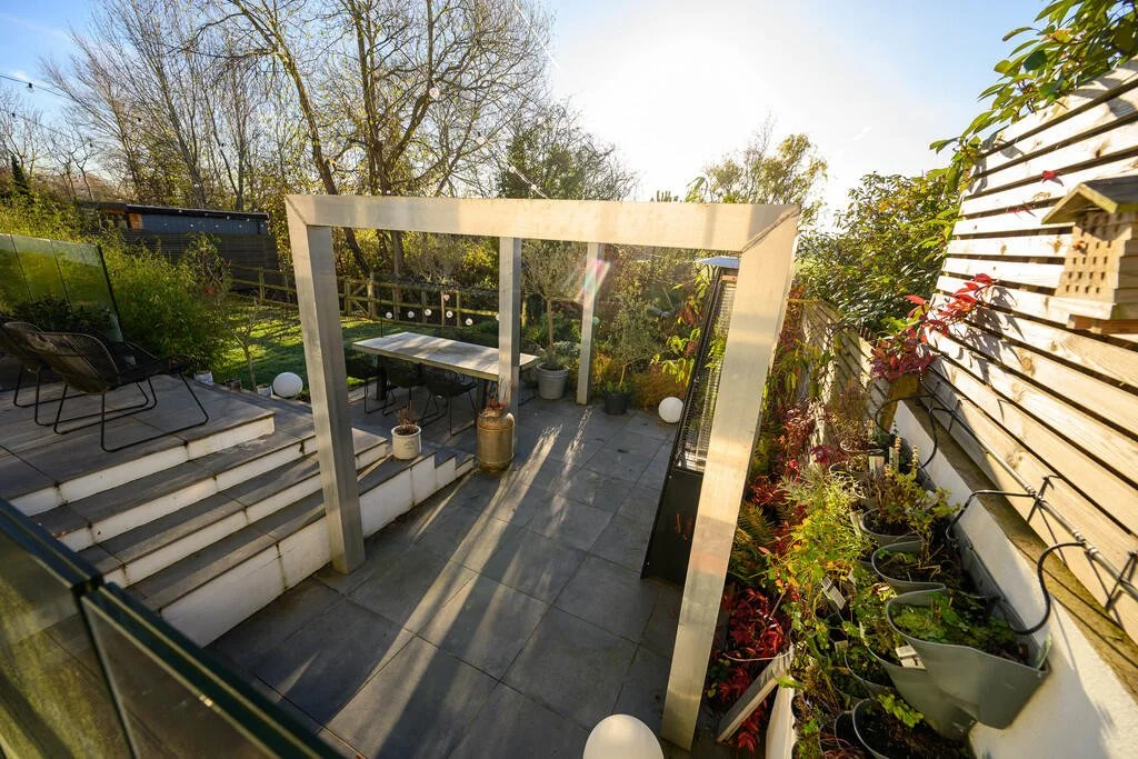 Outdoor patio area with potted plants, outdoor furniture, and a wooden fence, surrounded by trees and greenery, with sunlight shining.