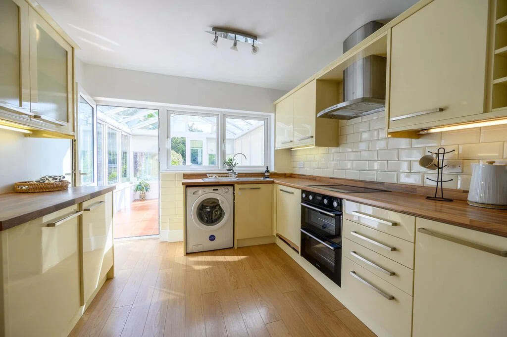 Merritt Estates. Bright kitchen with cream-colored cabinets, wooden countertops, white subway tile backsplash, oven, microwave, washing machine, and a small table with a kettle.