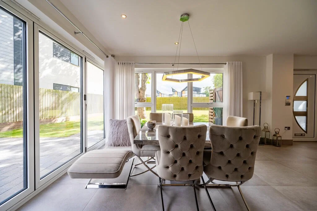 Merritt Estates. Modern dining room with a glass sliding door, a round dining table with six padded chairs, and a bright, cosy interior with white walls and curtains.
